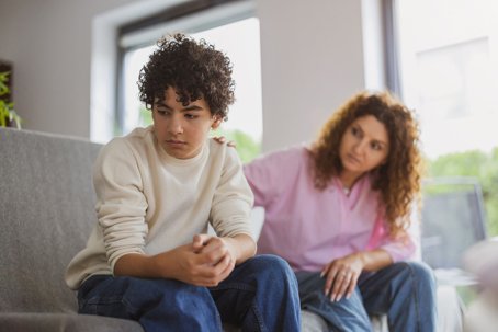 Upset boy sitting on sofa while his mother looking at him